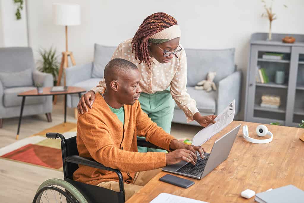 Portrait of African-American man using wheelchair working from home with wife looking over his shoulder, copy space