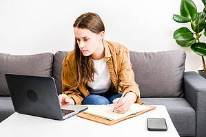 Focused young woman sit on sofa using laptop at home, looking at screen, chatting, reading or writing email, serious brunette female student doing homework, working on research project online concept